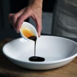 Person pouring dark vinegar into a white bowl on a cutting board
